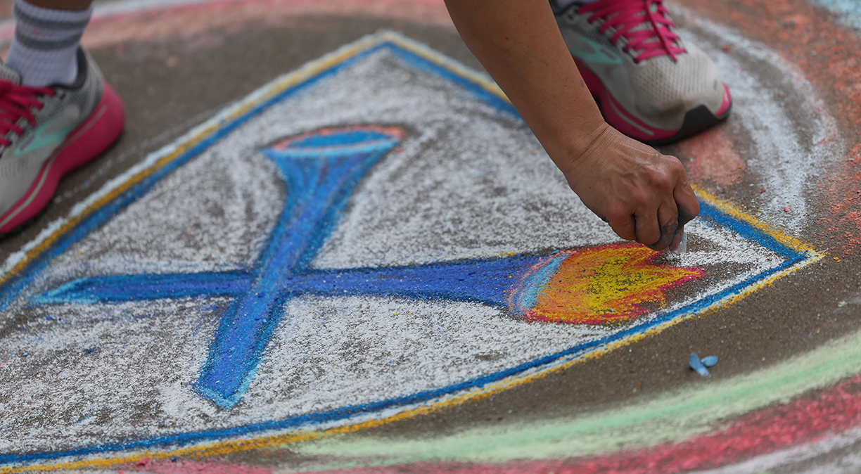 The hands of an artist are shown drawing the Emory logo in chalk on a paved sidewalk.